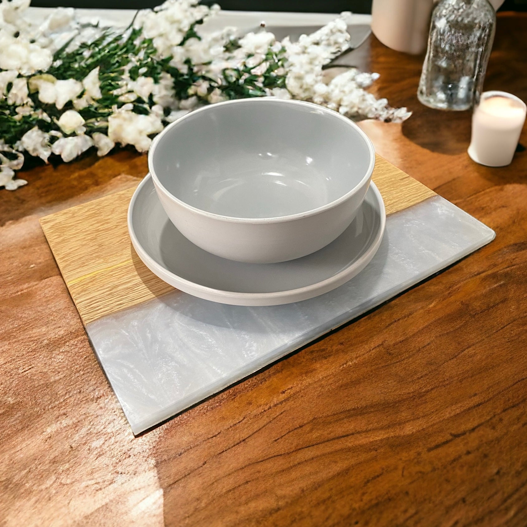 an oak and white resin placemat with a plate and bowl on it sat on a wooden dining table. white flowers and candles in the background
